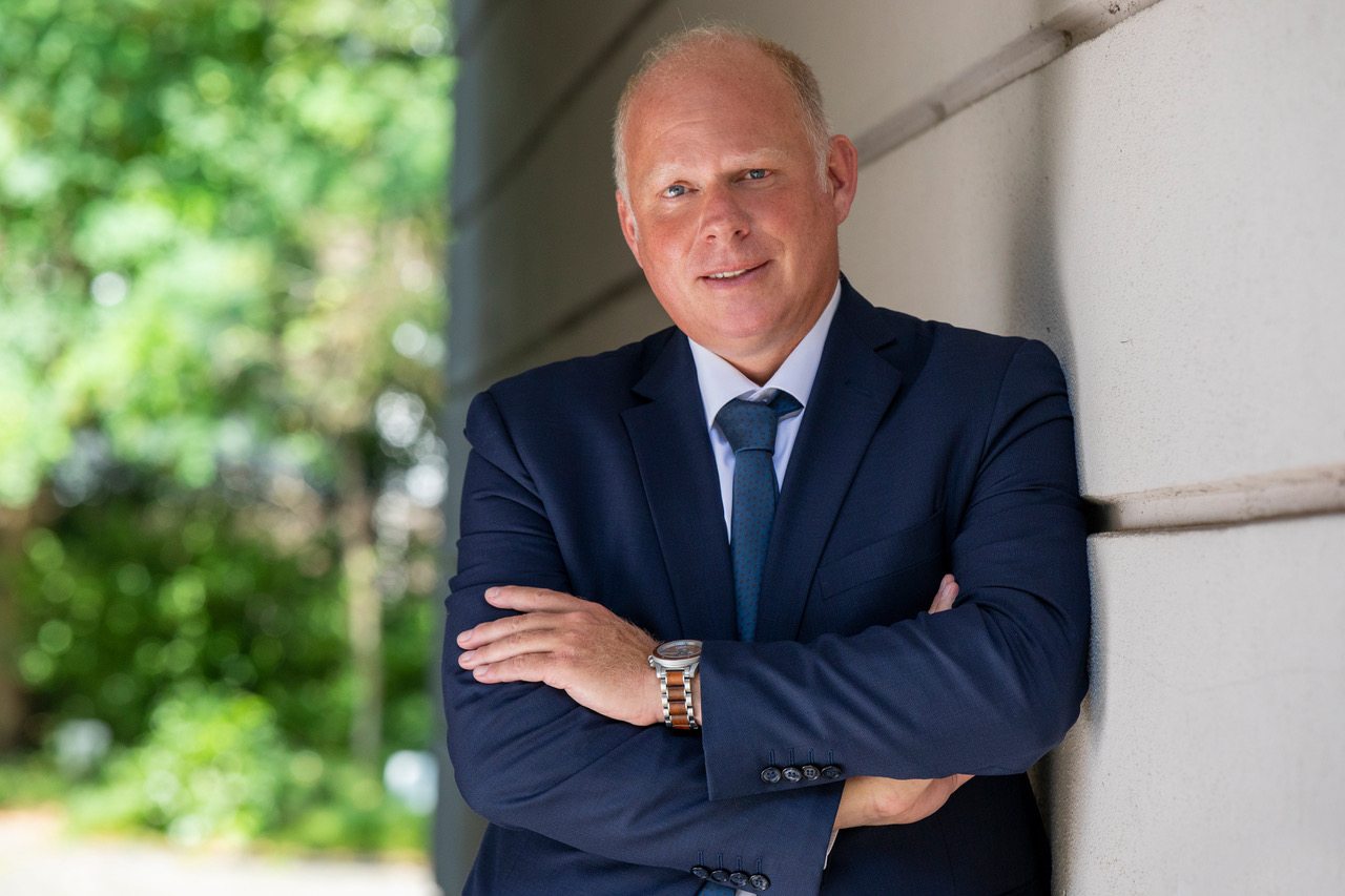 Ein lächelnder Mann in Anzug lehnt an einer Wand; hinter ihm ist unscharfe Natur zu sehen.